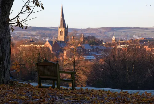 A view of Chesterfield Spire and town centre from Tapton park framed by a tree beside a park bench. The ground is covered in frost and fallen leaves.